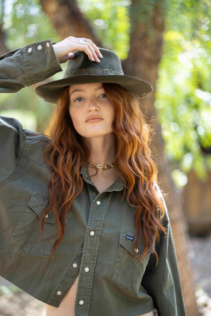 Model in an olive western shirt, hand on hat, under the trees